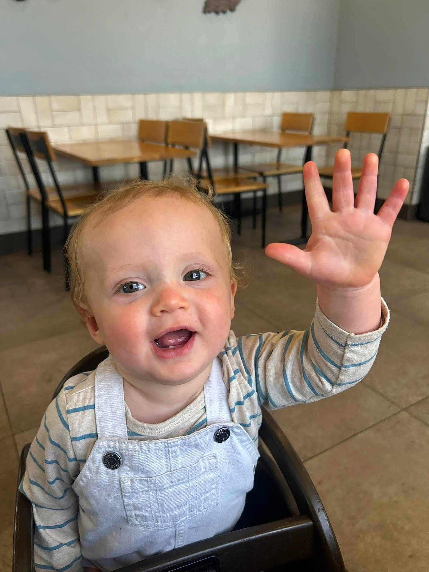 Colton joined the competition — help win amazing prizes! cafe, casual, child, curly_hair, cute, eyes, face, happy, high_chair, indoor, light_skin, mouth_open, overalls, person, smiling, striped_shirt, table, toddler, waving, wooden_chair