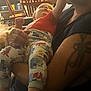 baby, adult, tattoo, pajamas, indoor, kitchen, furniture, wooden_chair, table, person, infant, holding, reaching, colorful_clothing, floor, home, arm, sitting, curious, casual