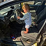 Colton is registered to the contest to win money with this photo: toddler, child, car_interior, steering_wheel, barefoot, overalls, curly_hair, dashboard, car_seat, door_open, sunlight, grass, electronic_device, floor_mat, vehicle, side_door, seat_belt, window, person, curiosity