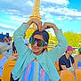 child, girl, sunglasses, heart_pose, eiffel_tower, landmark, outdoor, blue_sky, clouds, people, tourists, stadium_seats, colorful_clothing, pastel_colors, knit_sweater, smiling, daytime, trees, photography, happy