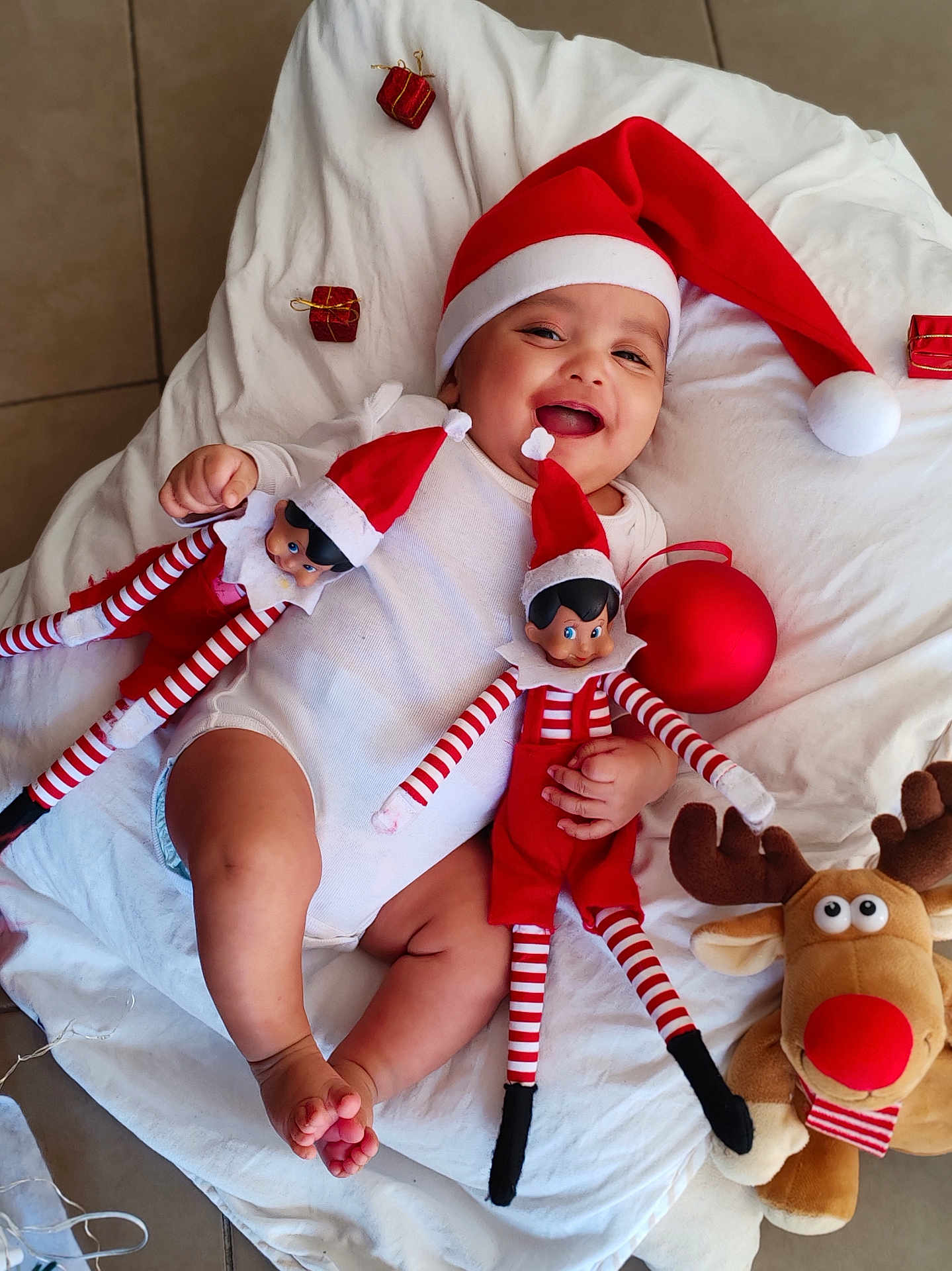 Evan David a rejoint le concours — aidez-le/la à gagner de superbes lots ! baby, child, santa_hat, smiling, toy, elf_doll, reindeer_plush, red, white_blanket, holiday, christmas, festive, cute, happy, indoor, floor, clothing, infant, playful, portrait