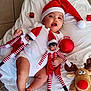 baby, infant, santa_hat, white_clothing, elf_doll, striped_clothing, red_and_white, plush_toy, reindeer_toy, christmas_decoration, red_ornament, blanket, floor_tiles, festive, holiday, cute, child, smiling, indoors, toy