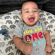 Xavier is registered to the contest to win money with this photo: baby, baby_girl_or_boy, child, closeup, clothing, cushion, cute, elephant_pattern, fabric, happy, indoor, infant, mickey_mouse, onesie, pacifier, person, portrait, relaxed, smiling, soft