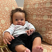 Ivanna is registered to the contest to win money with this photo: baby, infant, child, curly_hair, white_shirt, nike_logo, black_pants, couch, patterned_cushion, orange_cushion, toy, bare_feet, smile, indoors, comfortable, seated, portrait, cute, happy, person
