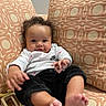 baby, infant, child, curly_hair, white_shirt, nike_logo, black_pants, couch, patterned_cushion, orange_cushion, toy, bare_feet, smile, indoors, comfortable, seated, portrait, cute, happy, person