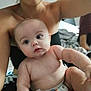 baby, infant, child, person, skin, face, arm, hand, torso, indoor, blurred_background, adult, holding, curious, wide_eyes, chubby_cheeks, soft_light, bed, blanket, casual