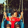 child, toddler, swing, playground, outdoor, sunlight, smiling, happy, ropes, blue_swing, red_safety_bar, barefoot, grass, trees, fence, summer, play, person, casual_clothing, daylight