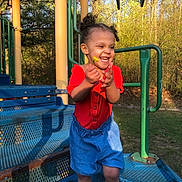 Alina is registered to the contest to win money with this photo: child, toddler, playground, flower, smile, happy, outdoor, curly_hair, red_clothing, blue_shorts, sunlight, trees, grass, metal_structure, play_equipment, person, nature, summer, joy, cute