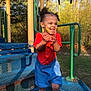 child, toddler, playground, flower, smile, happy, outdoor, curly_hair, red_clothing, blue_shorts, sunlight, trees, grass, metal_structure, play_equipment, person, nature, summer, joy, cute