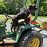 dog, tractor, outdoor, sunny, parked_vehicle, farm_equipment, agriculture, machinery, green_tractor, large_tire, yellow_rim, dog_collar, open_mouth, panting, tongue, black_and_white_dog, trees, grass, gravel, playful