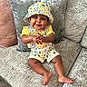 baby, child, smiling, clapping, yellow_clothing, hat, floral_pattern, couch, cushion, indoor, happy, cute, sitting, infant, portrait, plush, comfort, home, person, bright