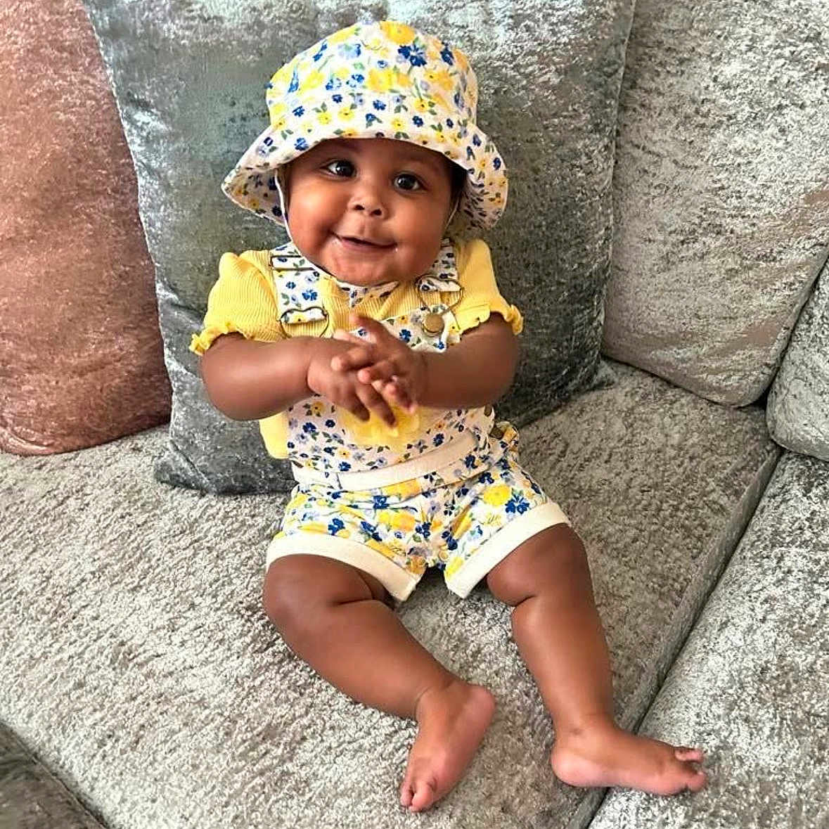 Zarah is registered to the contest to win money with this photo: baby, child, smiling, clapping, yellow_clothing, hat, floral_pattern, couch, cushion, indoor, happy, cute, sitting, infant, portrait, plush, comfort, home, person, bright