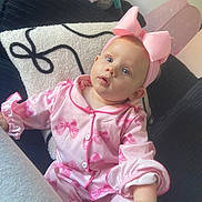 River is registered to the contest to win money with this photo: baby, child, pink, pajamas, bow, headband, couch, cushion, indoor, person, portrait, infant, clothing, cute, seated, looking_up, soft_lighting, home, relaxing, expression