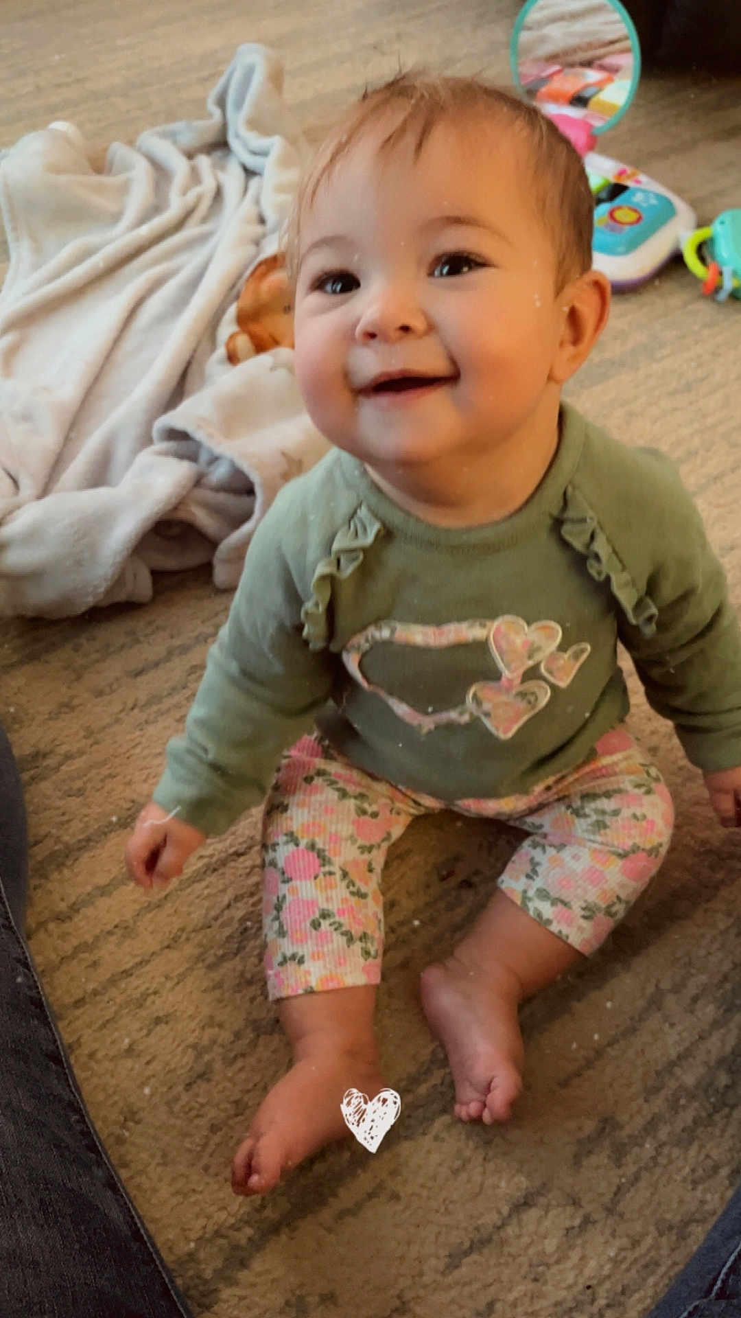 Chloe is registered to the contest to win money with this photo: baby, child, smiling, sitting, green_sweater, floral_pants, carpet, toys, blanket, indoors, cute, happy, face, feet, person, toddler, playtime, home, infant, warm