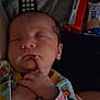 baby, sleeping, infant, blanket, onesie, colorful_clothing, hand, face, remote_control, snacks, dim_light, indoors, cute, peaceful, soft_texture, newborn, portrait, resting, child, cozy