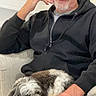 man, elderly, person, dog, small_dog, pet, companion, portrait, indoors, couch, cushion, blue_cushion, black_hoodie, white_hair, beard, sitting, relaxing, cozy, home, photography