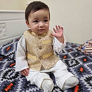Aryan is registered to the contest to win money with this photo: toddler, child, sitting, waving, golden_vest, white_clothing, bedspread, patterned_bedspread, indoors, cute, person, portrait, young_child, smiling, hand_raised, happy, face, short_hair, traditional_clothing, baby