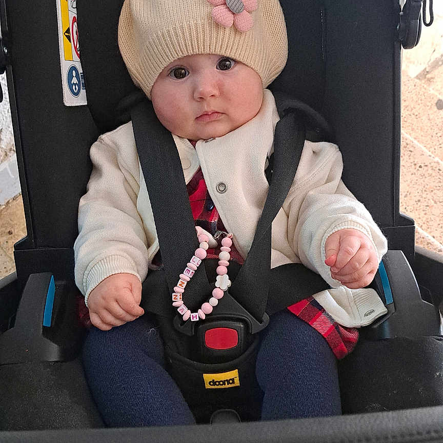 Jennah a rejoint le concours — aidez-le/la à gagner de superbes lots ! baby, baby_accessory, beaded_bracelet, blue_pants, car_seat, child, clothing, cute, face, floor, flower, hat, indoor, infant, jacket, person, pink_shoe, portrait, seatbelt, sitting