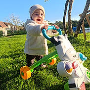 Emeraude a rejoint le concours — aidez-le/la à gagner de superbes lots ! toddler, child, outdoor, grass, walker_toy, toy, cow_toy, hat, jacket, sunshine, blue_sky, trees, farm_buildings, greenery, playing, smiling, happy, person, daylight, nature