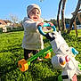 toddler, child, outdoor, grass, walker_toy, toy, cow_toy, hat, jacket, sunshine, blue_sky, trees, farm_buildings, greenery, playing, smiling, happy, person, daylight, nature