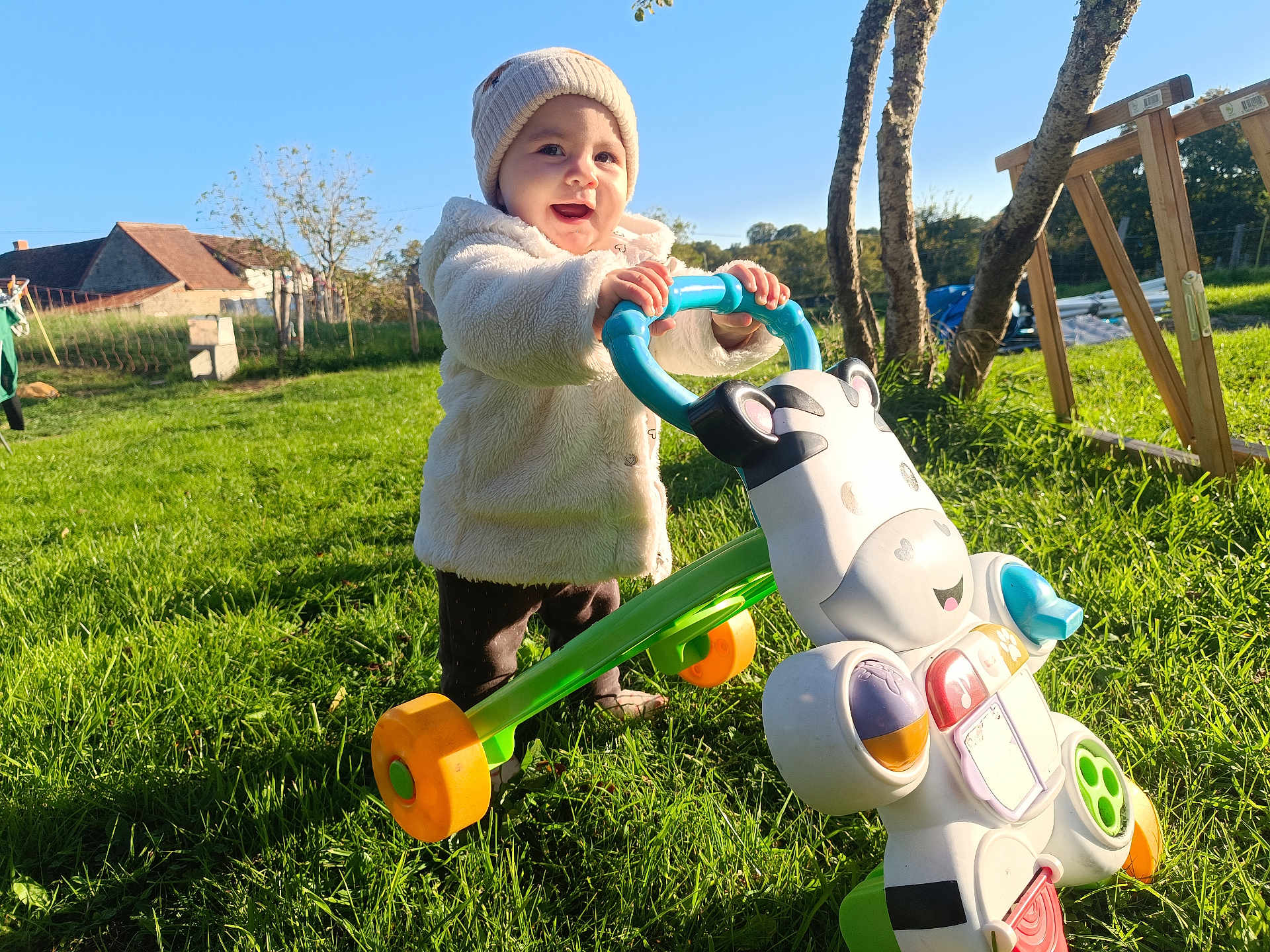 Emeraude a rejoint le concours — aidez-le/la à gagner de superbes lots ! toddler, child, outdoor, grass, walker_toy, toy, cow_toy, hat, jacket, sunshine, blue_sky, trees, farm_buildings, greenery, playing, smiling, happy, person, daylight, nature