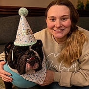 Sir Winston Dean is registered to the contest to win money with this photo: woman, dog, bulldog, party_hat, bandana, smiling, indoor, couch, celebration, casual_clothing, long_hair, portrait, pet, companion, happy, cozy, home, ripped_jeans, hand, friendship