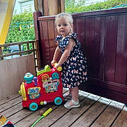 Maddy participe au concours pour gagner de l'argent avec cette photo : toddler, child, toy_train, dress, butterfly_pattern, wooden_floor, outdoor, porch, fence, greenery, shoes, play, colorful_toy, person, standing, summer, daylight, curtain, plastic_toy, happy