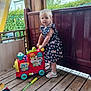 toddler, child, toy_train, dress, butterfly_pattern, wooden_floor, outdoor, porch, fence, greenery, shoes, play, colorful_toy, person, standing, summer, daylight, curtain, plastic_toy, happy