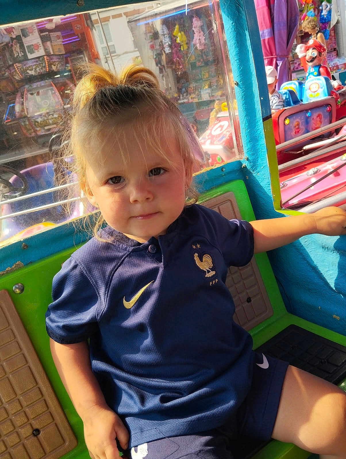 Maddy a rejoint le concours — aidez-le/la à gagner de superbes lots ! toddler, child, amusement_ride, carnival, blonde_hair, pony_tail, blue_shirt, sportswear, indoor, colorful, smiling, seat, toy_ride, face, person, daylight, playful, entertainment, fun, background