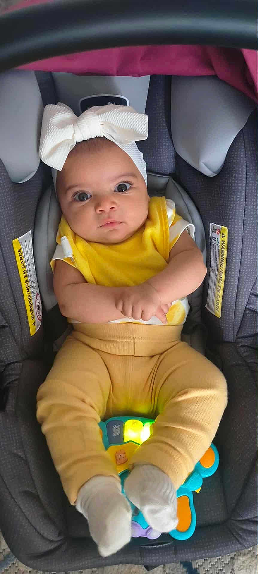 Amna joined the competition — help win amazing prizes! baby, child, car_seat, yellow_clothing, headband, toy, light_up_toy, socks, infant, cute, indoors, seatbelt, portrait, looking_up, hands, feet, playful, expression, sitting, comfort