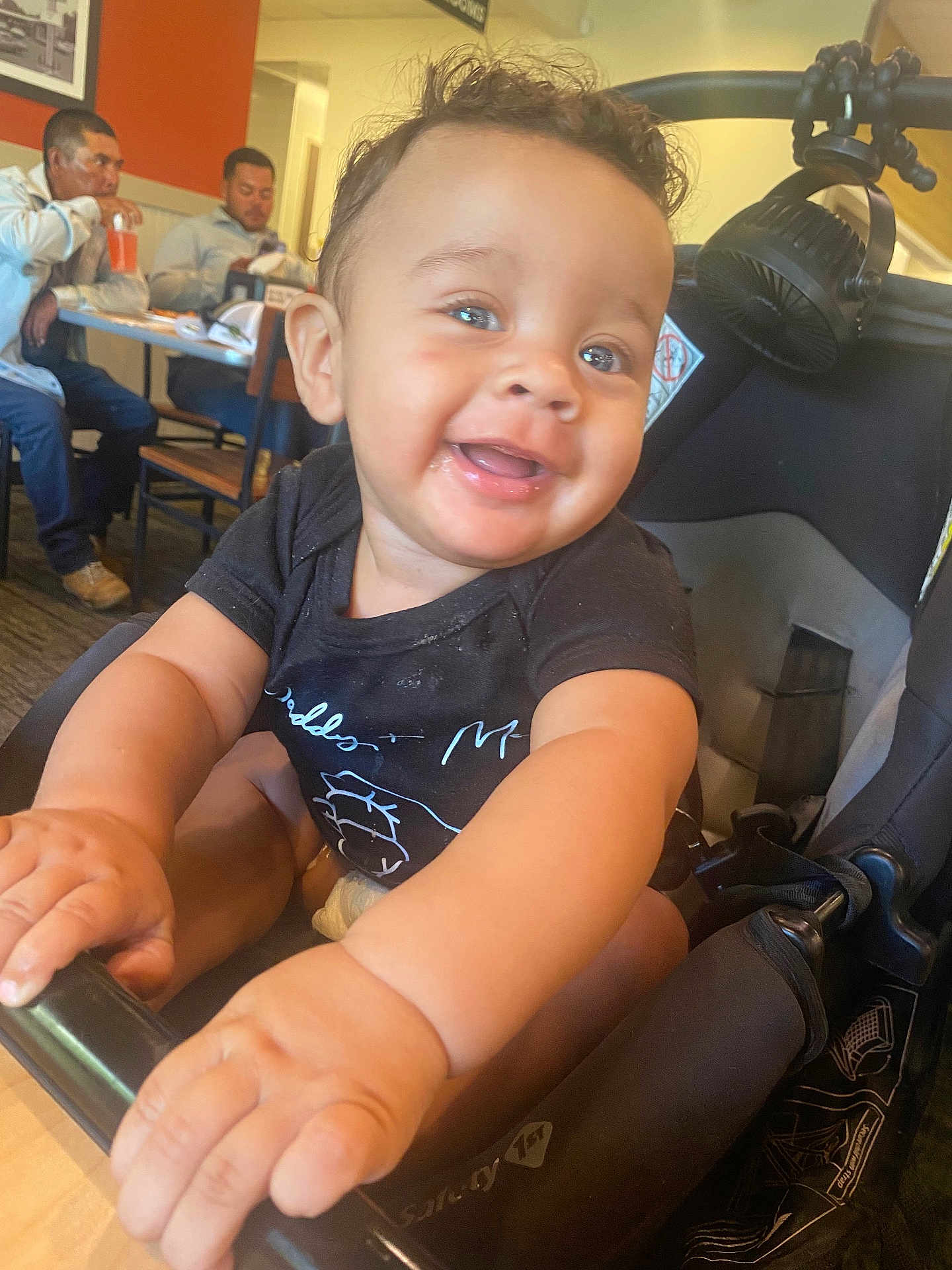 Tre is registered to the contest to win money with this photo: baby, smile, hands, stroller, indoor, restaurant, people, man, table, chair, drink, clothing, face, person, happy, child, casual, background, light, curly_hair