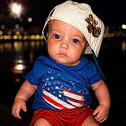 Ezekiel is registered to the contest to win money with this photo: baby, child, blue_shirt, red_shorts, white_cap, hat, face, portrait, night, fireworks, lights, outdoor, people, american_flag, expression, sitting, cute, fashion, summer, photography