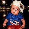 baby, child, blue_shirt, red_shorts, white_cap, hat, face, portrait, night, fireworks, lights, outdoor, people, american_flag, expression, sitting, cute, fashion, summer, photography