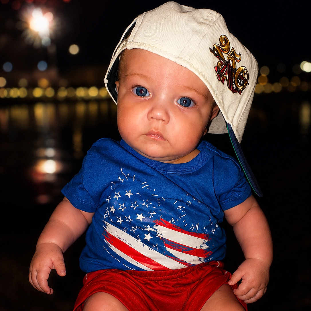 Ezekiel is registered to the contest to win money with this photo: baby, child, blue_shirt, red_shorts, white_cap, hat, face, portrait, night, fireworks, lights, outdoor, people, american_flag, expression, sitting, cute, fashion, summer, photography