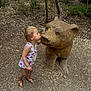 child, toddler, dress, sandals, wooden_bear, sculpture, forest, trees, nature, outdoor, ground, rocks, plants, cute, interaction, playful, daylight, person, kissing, wood