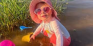 Mya a rejoint le concours — aidez-le/la à gagner de superbes lots ! child, toddler, water, lake, sunglasses, hat, playing, plastic_toys, shallow_water, reeds, outdoor, sunny, summer, splashing, pink, yellow, blue, nature, happy, fun