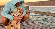 Bolt a rejoint le concours — aidez-le/la à gagner de superbes lots ! person, dog, puppy, pet, hoodie, glasses, cuddle, outdoor, seaside, pier, dock, ocean, water, sunset, sky, wooden_deck, railing, leash, harness, portrait