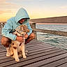 person, dog, puppy, pet, hoodie, glasses, cuddle, outdoor, seaside, pier, dock, ocean, water, sunset, sky, wooden_deck, railing, leash, harness, portrait