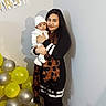 baby, balloons, birthday, celebration, child, family, hat, home, indoors, infant, mother, party_banner, portrait, scarf, silver_balloons, smiling, standing, winter_clothing, woman, yellow_balloons