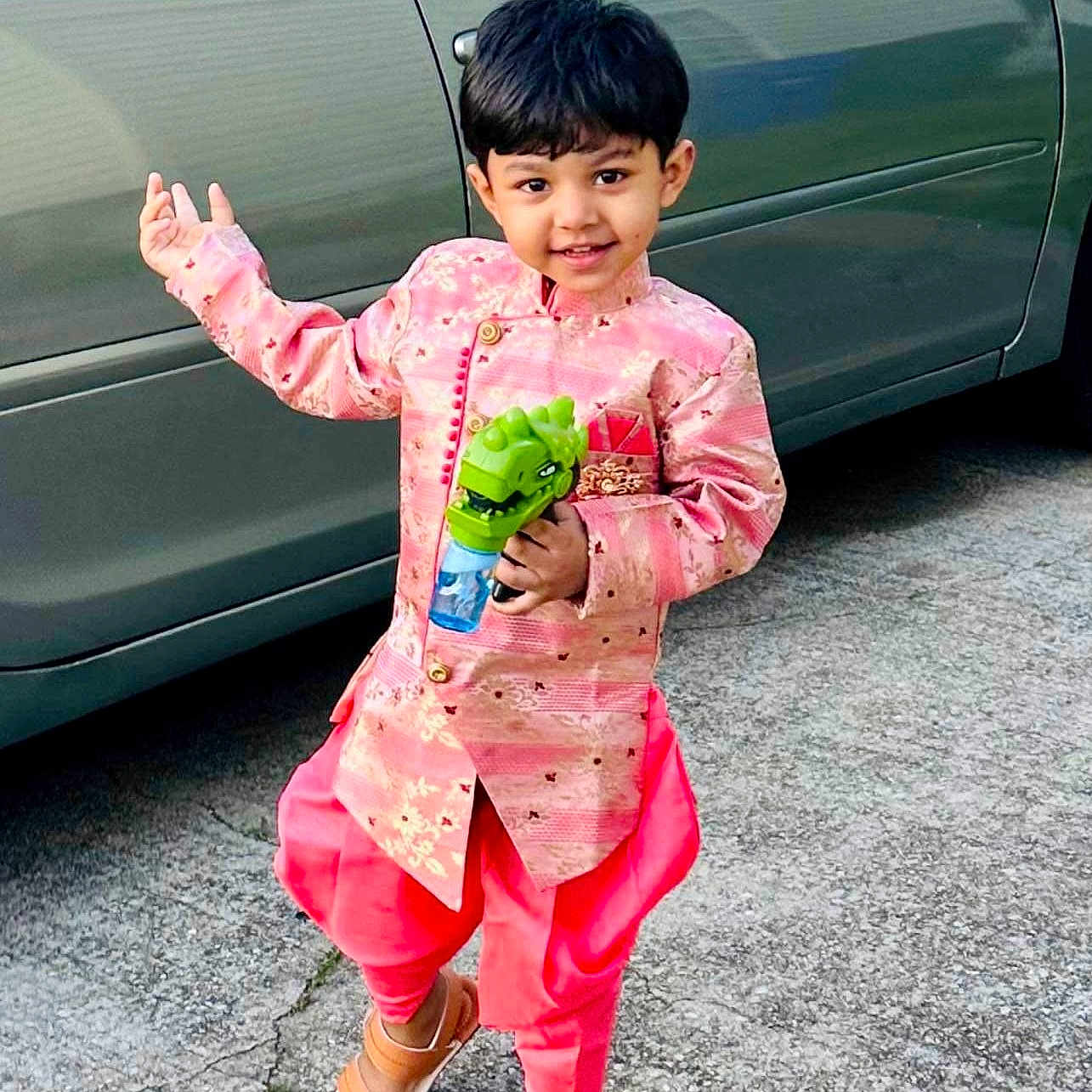 Arish is registered to the contest to win money with this photo: boy, car, child, daylight, driveway, fashion, formalwear, full_body, green_toy, outdoor, pavement, pink_outfit, portrait, sandals, short_hair, smiling, standing, toy, traditional_clothing, vehicle
