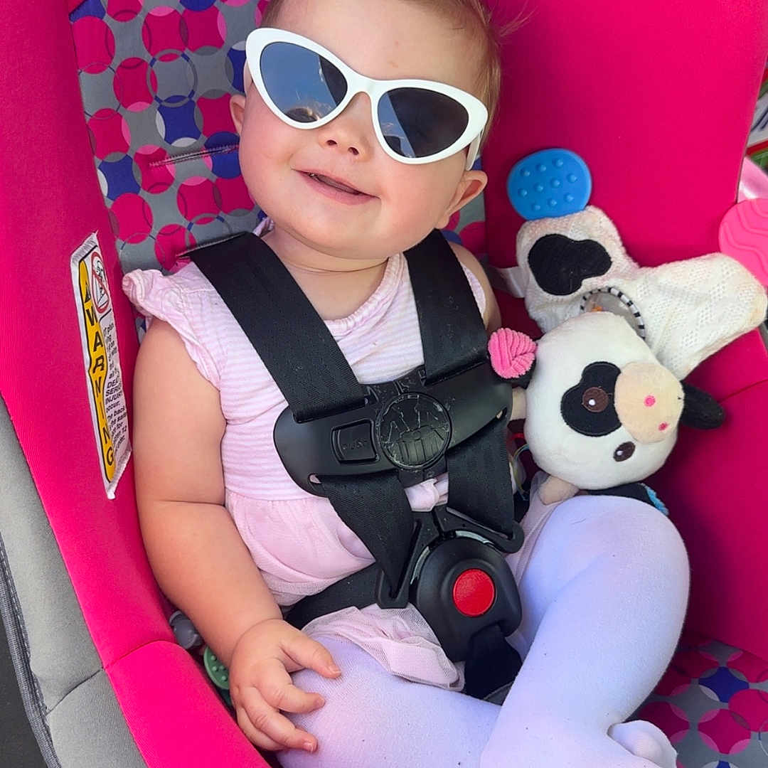 Willow is registered to the contest to win money with this photo: accessory, baby, car_seat, child, clothing, cow, cute, fashion, happy, indoor, infant, person, pink, plush_toy, portrait, safety_harness, seatbelt, smile, sunglasses, toy