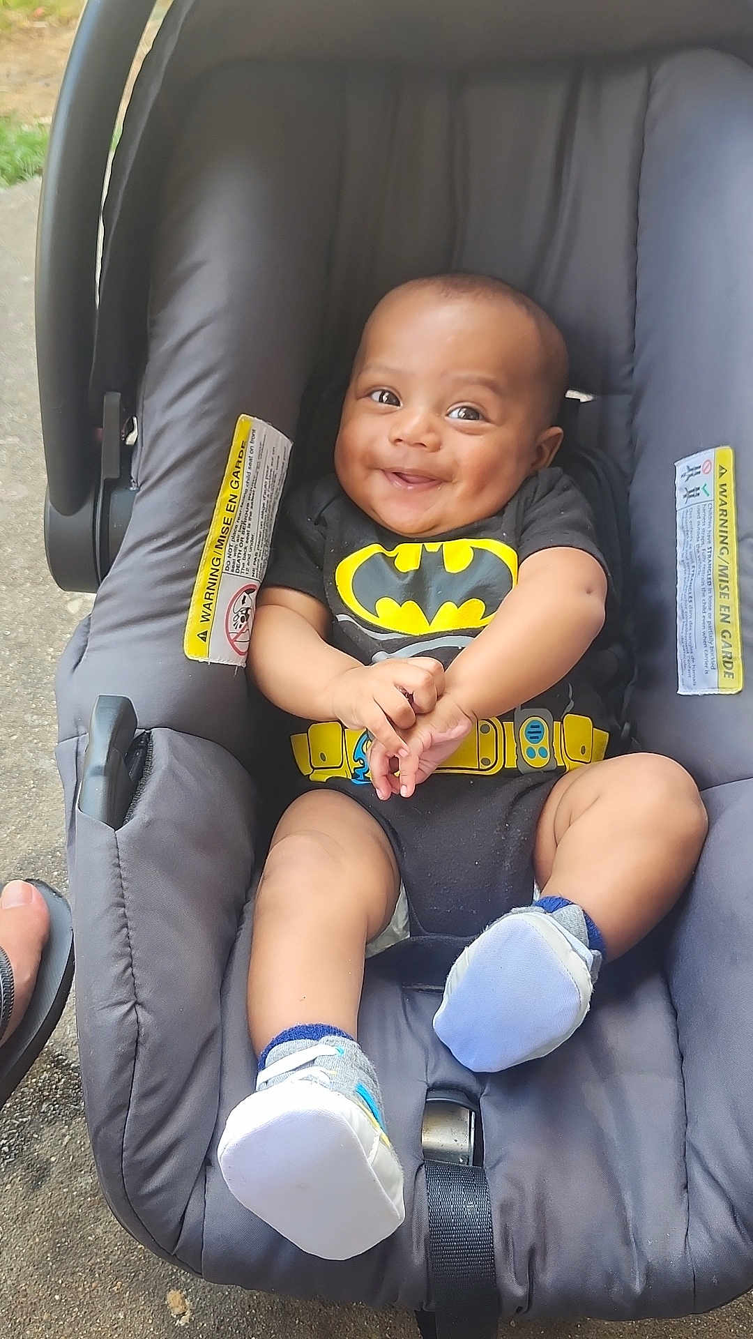Kaison is registered to the contest to win money with this photo: baby, infant, car_seat, smile, batman, onesie, dimples, footwear, socks, hands, outdoor, grass, concrete, happy, child, person, seat_belt, foot, apparel, cute