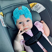 Cora is registered to the contest to win money with this photo: baby, child, car_seat, blue_bow, headband, pink_clothing, toy, safety_strap, warning_label, infant, cute, portrait, hands, face, seat, child_safety, car_interior, expression, young, secure