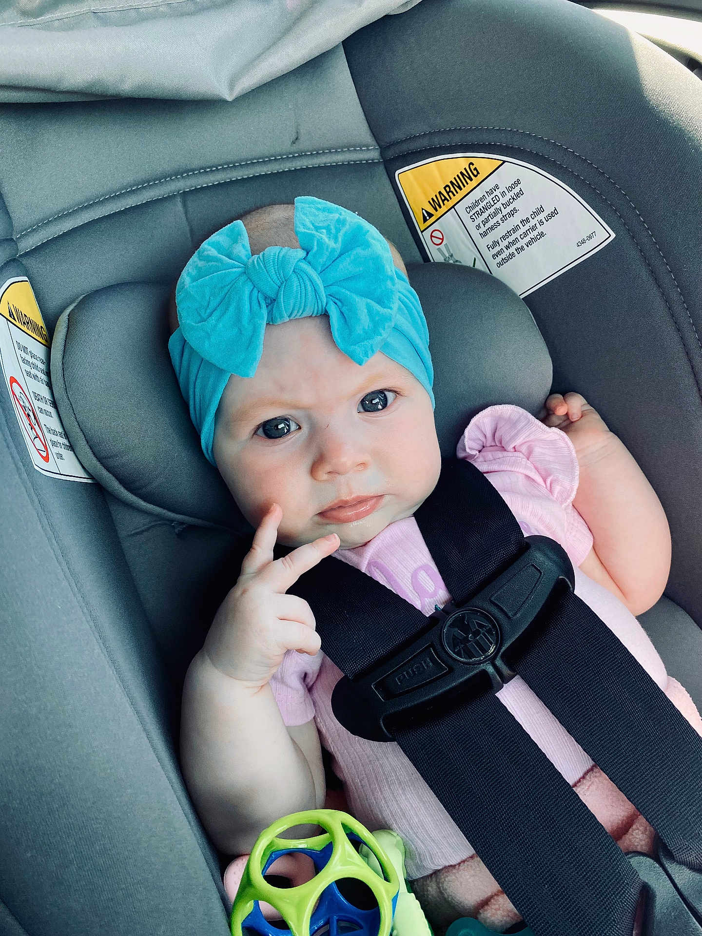 Cora is registered to the contest to win money with this photo: baby, child, car_seat, blue_bow, headband, pink_clothing, toy, safety_strap, warning_label, infant, cute, portrait, hands, face, seat, child_safety, car_interior, expression, young, secure