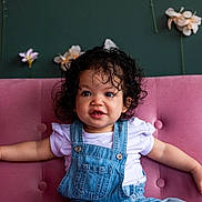 Alani is registered to the contest to win money with this photo: toddler, child, curly_hair, denim_overalls, white_shirt, pink_couch, green_wall, flowers, sitting, indoor, portrait, cute, happy, baby, person, smiling, furniture, casual_clothing, decor, young_child