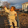 child, toddler, playground, spring_rocker, wood_chips, pointing, white_jacket, pants, sneakers, sitting, surprised_expression, golden_hour, sunlight, outdoor, fence, residential, portrait, toy, play, babyface