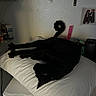 black_cat, cat, pillow, white_pillow, stretched_out, tail_curl, indoor, furniture, table, shelf, poster, cartoon, dim_lighting, domestic, pet, relaxed, sleeping, animal, cozy, resting