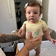 Diana participe au concours pour gagner de l'argent avec cette photo : baby, child, infant, tattoo, hands, bow, shirt, standing, changing_table, indoor, person, expression, foot, skin, hair, furniture, tattooed_arms, support, room, curious