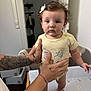 baby, child, infant, tattoo, hands, bow, shirt, standing, changing_table, indoor, person, expression, foot, skin, hair, furniture, tattooed_arms, support, room, curious