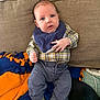 baby, infant, plaid_shirt, blue_eyes, green_socks, navy_pants, bib, couch, blanket, indoor, child, person, cute, young, seated, cozy, comfortable, soft_fabric, expression, casual_clothing